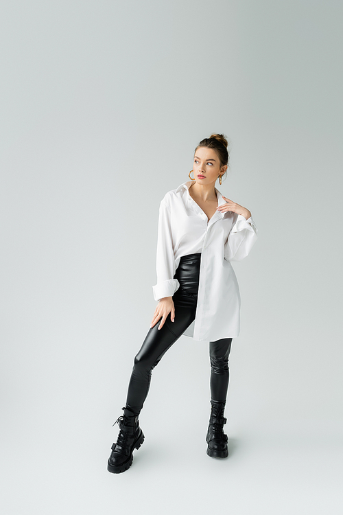 full length of trendy woman in oversize shirt and tight pants with rough boots looking away on grey background