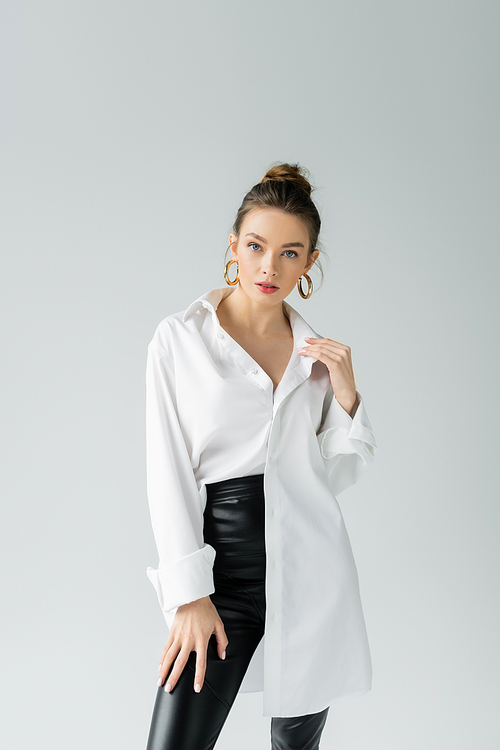 fashionable woman in white oversize shirt and golden earrings looking at camera isolated on grey