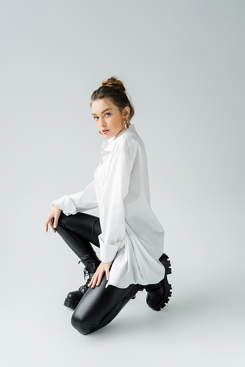 full length of young woman in white shirt and black rough boots looking at camera while posing on grey background