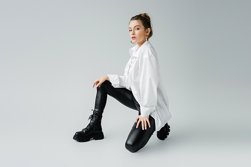 full length of trendy model in black latex pants and oversize shirt looking at camera on grey background