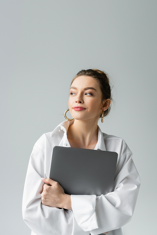 happy young woman in white shirt holding laptop and looking away isolated on grey