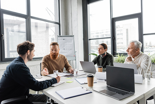 Bearded businessman talking to colleagues near devices and papers during training in office