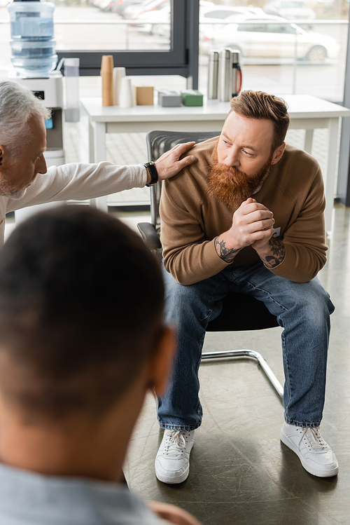 Middle aged man calming tattooed person with alcohol addiction during therapy in rehab center