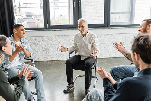 Positive mature man with alcohol addiction sitting in circle while multiethnic people applauding in rehab center