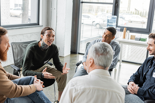 Cheerful tattooed man with alcohol addiction talking to interracial group during therapy in rehab center