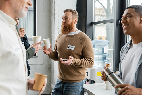 Bearded man with paper cup and smartphone talking to people during alcoholics meeting in recovery center