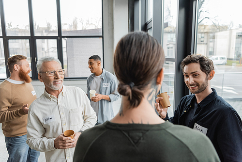 Positive people with paper cups looking at blurred tattooed man during alcoholics meeting in rehab center