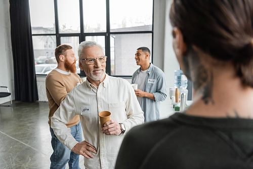 Smiling mature man with paper cup and name sticker spending time during alcoholics meeting in rehab center