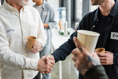 Cropped view of people holding paper cups and shaking hands during alcoholics meeting in rehab center