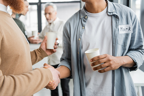 Interracial men with paper cups shaking hands during alcoholics meeting in rehab center