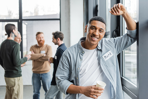 Smiling african american man holding paper cup near blurred group during alcoholics meeting in rehab center