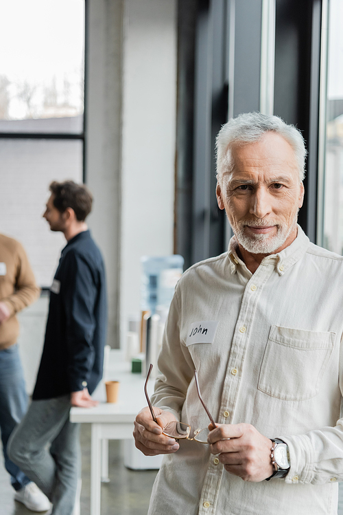 Middle aged man holding eyeglasses and looking at camera during alcoholics meeting in rehab center