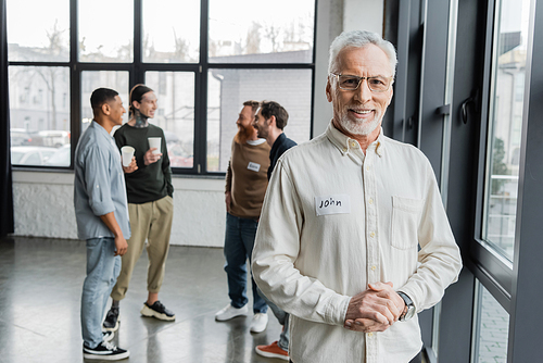 Smiling middle aged man looking at camera near blurred group during alcoholics meeting in rehab center