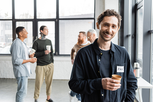 Smiling man holding paper cup and looking at camera during alcoholics meeting in rehab center