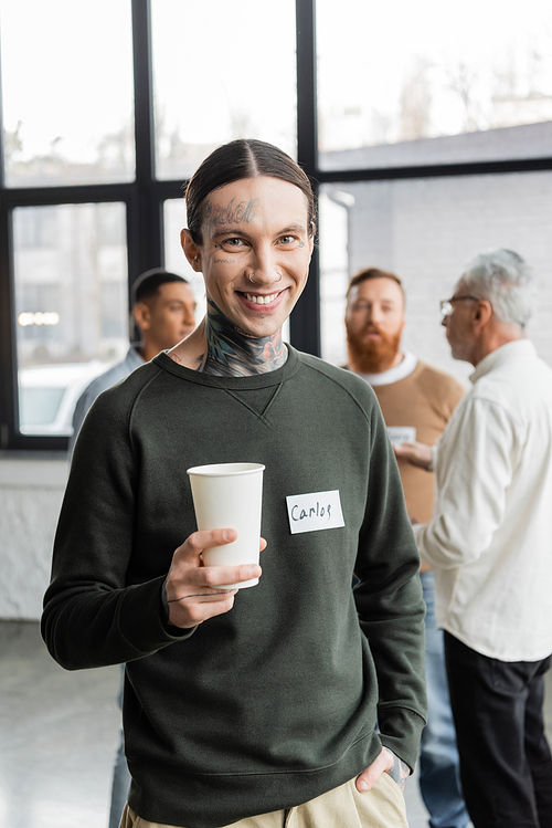 Smiling tattooed man holding paper cup and looking at camera during alcoholics meeting in recovery center