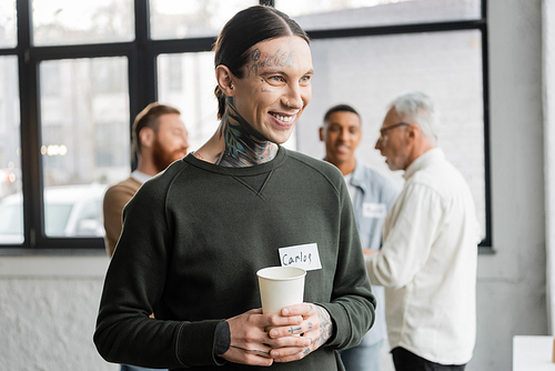 Positive tattooed man holding paper cup during meeting of alcoholics in recovery center