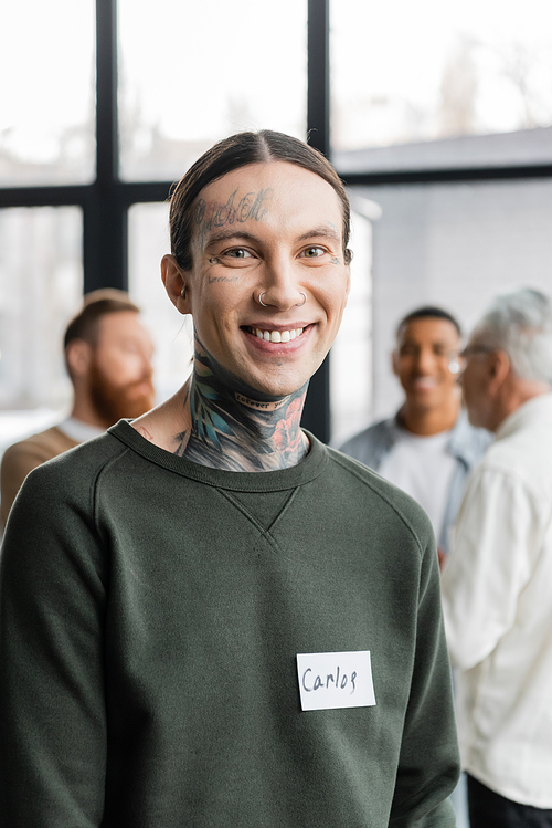 Portrait of smiling tattooed man with name sticker looking at camera during aa meeting in rehab center