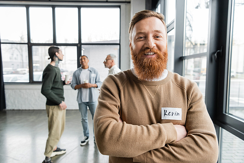 Cheerful bearded man crossing arms and looking at camera during alcoholics meeting in rehab center