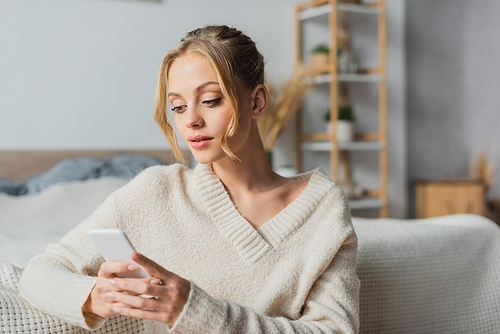 young blonde woman in knitted sweater chatting on smartphone in modern bedroom