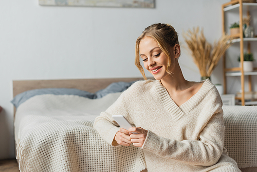 cheerful young woman in sweater using smartphone in modern bedroom