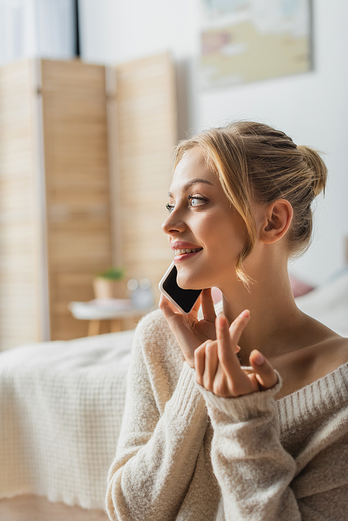 cheerful young woman in sweater talking on smartphone in modern bedroom