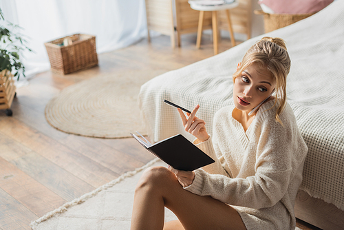 young blonde woman in sweater talking on smartphone and holding pen near notebook in bedroom