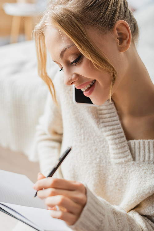 smiling woman in sweater talking on smartphone and taking notes in notebook