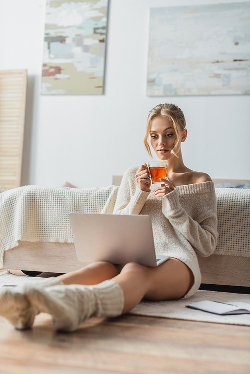 blonde woman holding glass cup of tea while looking at laptop in modern apartment