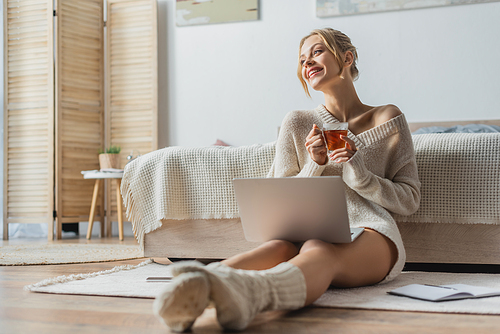 happy woman holding glass cup of tea while sitting with laptop in modern apartment