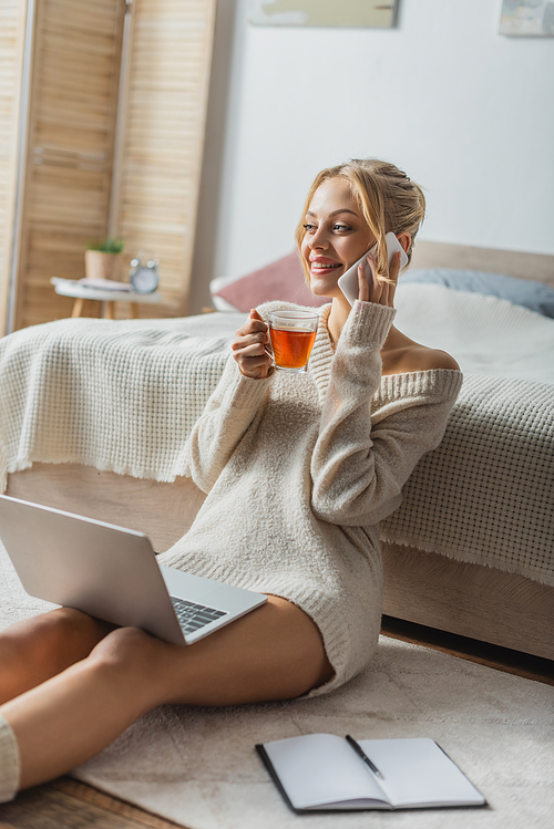 happy woman holding glass cup of tea while sitting with laptop and talking on smartphone in modern apartment