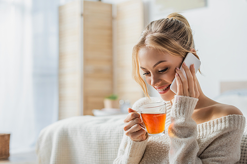 happy woman holding glass cup of tea while talking on smartphone in modern apartment