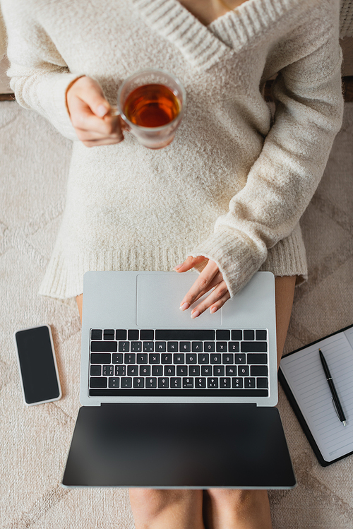 top view of cropped woman holding glass cup with tea while using laptop near smartphone with blank screen