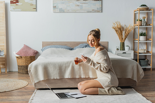 blonde woman holding glass cup with tea and using smartphone near laptop on carpet