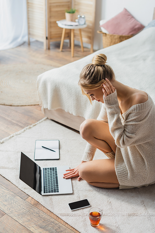 blonde woman in sweater using laptop near mobile phone and cup of tea on carpet in bedroom