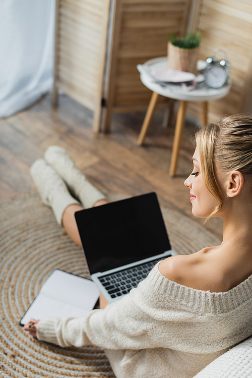 charming woman in sweater and socks using laptop in modern apartment