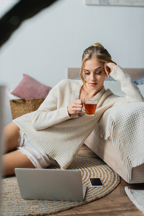 young blonde woman in sweater holding glass cup with tea and looking at laptop in bedroom
