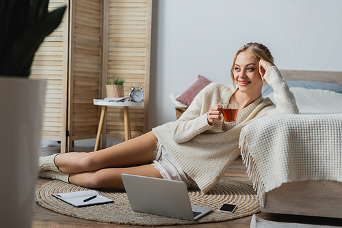 smiling blonde woman in sweater holding glass cup with tea while sitting near gadgets in bedroom