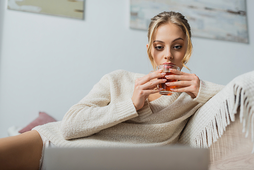 blonde woman in sweater holding glass cup with tea and looking at blurred laptop in bedroom