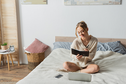 cheerful blonde woman taking notes near gadgets on bed in modern apartment