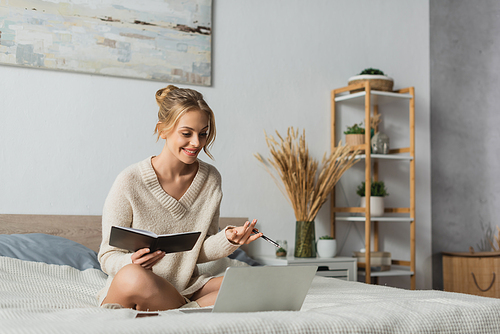 cheerful young woman holding notebook and pen while watching webinar on laptop in bedroom
