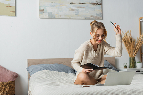 cheerful young woman holding notebook and pen while studying online in bedroom