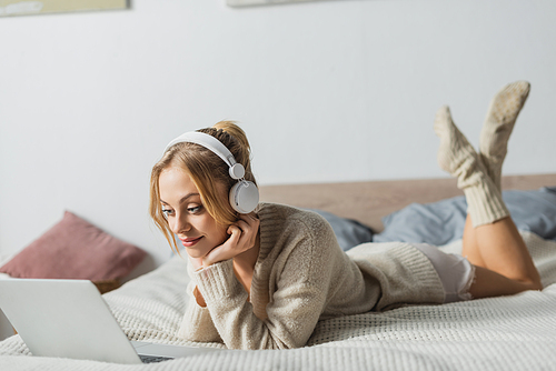 young blonde woman in wireless headphones watching movie on laptop in bedroom