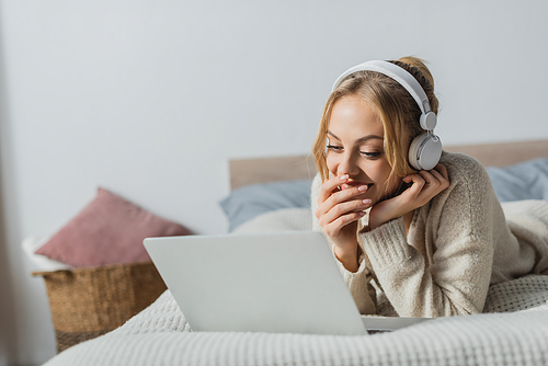 cheerful woman in wireless headphones watching comedy movie on laptop in bedroom