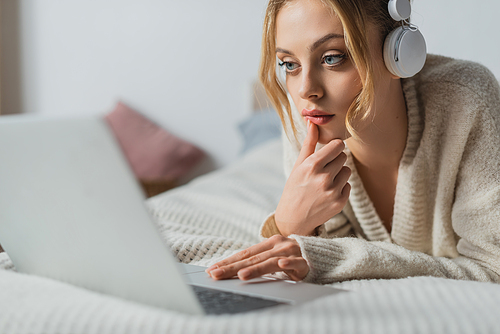 focused woman in wireless headphones using laptop in bedroom