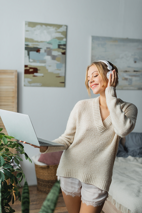 happy young woman in wireless headphones listening music and holding laptop in modern bedroom