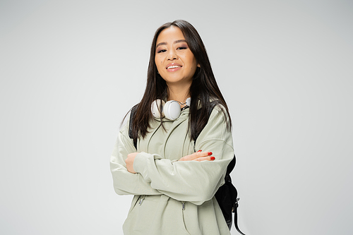 pretty asian student with wireless headphones and backpack posing with crossed arms isolated on grey