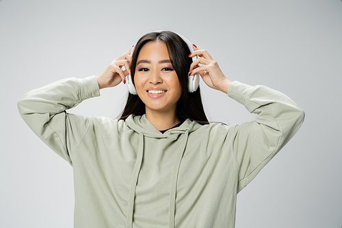 joyful asian woman in hoodie adjusting wireless headphones and looking at camera isolated on grey