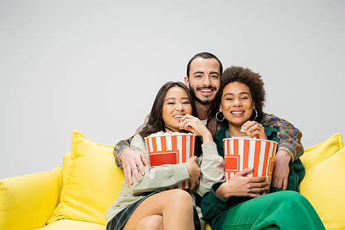 cheerful bearded man embracing interracial women eating popcorn and watching movie on yellow couch isolated on grey