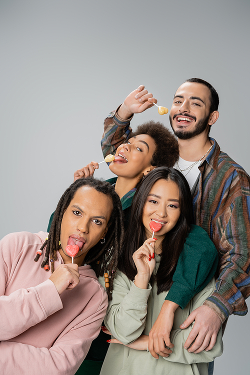 cheerful multiethnic friends in trendy clothes eating sweet lollipops isolated on grey