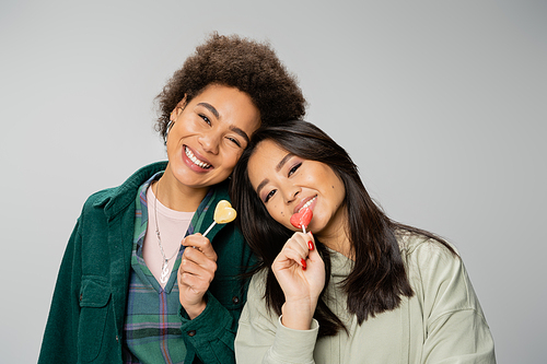 young and trendy multiethnic women with sweet lollipops looking at camera isolated on grey